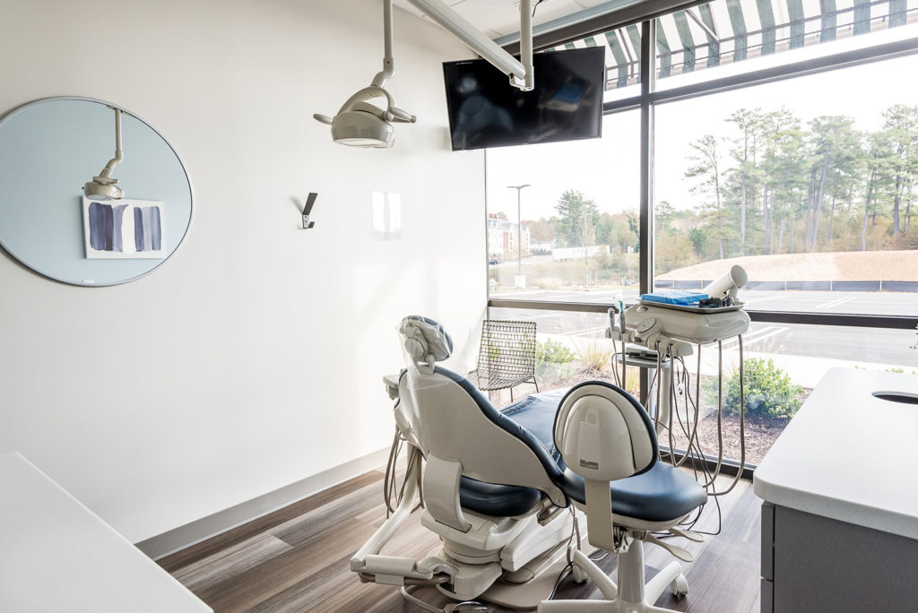 Modern dental treatment room with patient chair, mounted TV, and large windows providing natural light