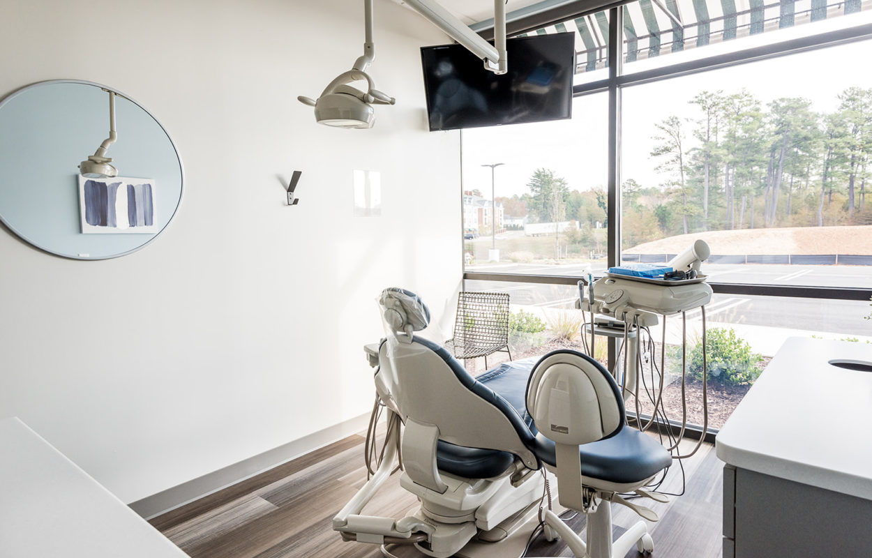 Modern dental treatment room with patient chair, mounted TV, and large windows providing natural light
